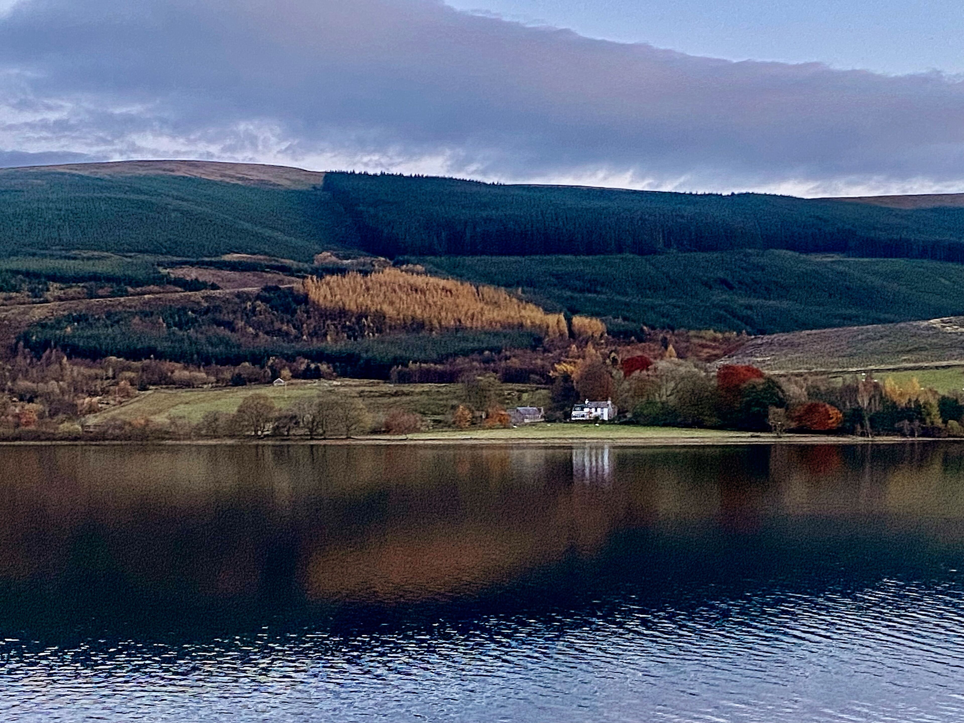 Un lac avec une maison sur la rive, entouré de collines et d’arbres.
