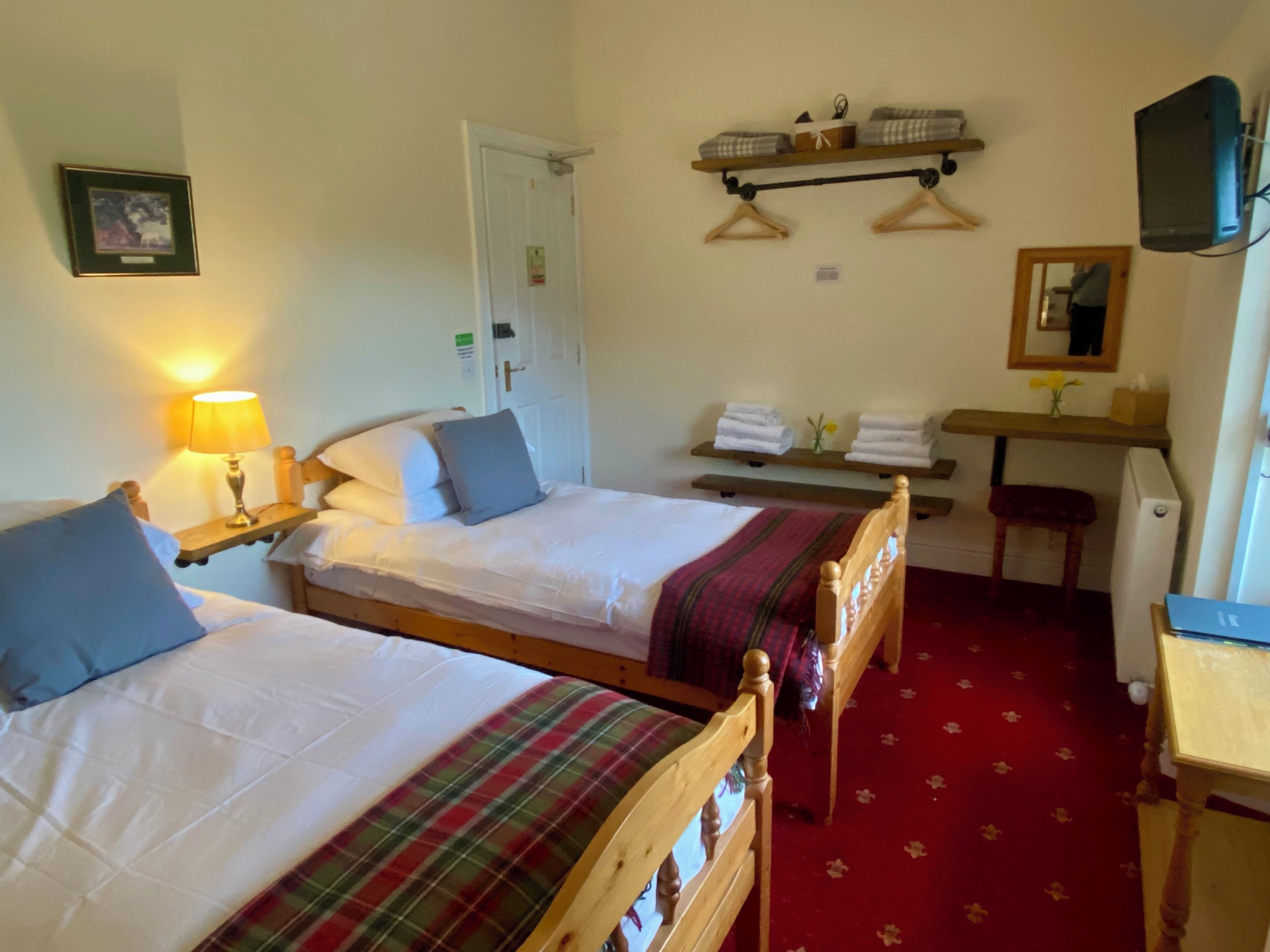 A hotel room with two single beds, a wooden desk, a chair, a television, and a shelf with towels.