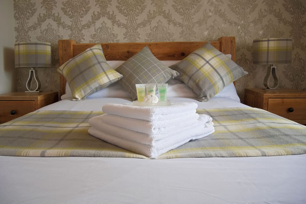 A neatly made bed with a plaid bedspread, two pillows, and a stack of white towels. There are two bedside tables with lamps and a decorative vase on one.