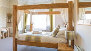 Una cama con dosel de madera, una mesita de noche, una lámpara y una silla en una habitación con papel tapiz estampado y un radiador.