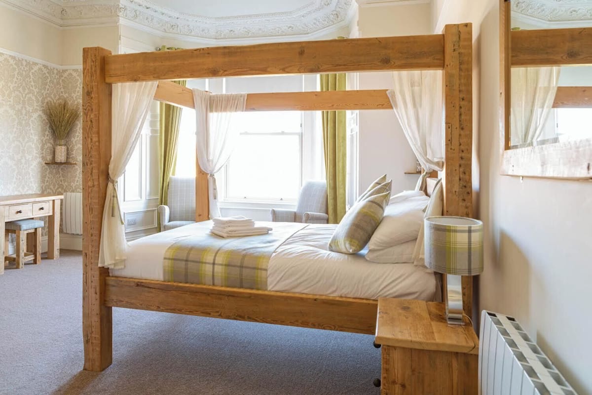 A wooden four-poster bed with a canopy, a bedside table, a lamp, and a chair in a room with patterned wallpaper and a radiator.