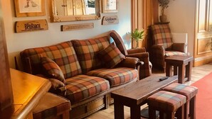 Una sala de estar acogedora con un sofá a cuadros, una mesa de centro de madera y otomanas a juego. Se observa también una estantería con diversos objetos y una planta en maceta.