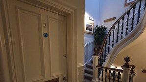 Un pasillo con una puerta blanca que dice 'The Dalmore', una escalera con barandilla de madera y un cuadro en la pared.
