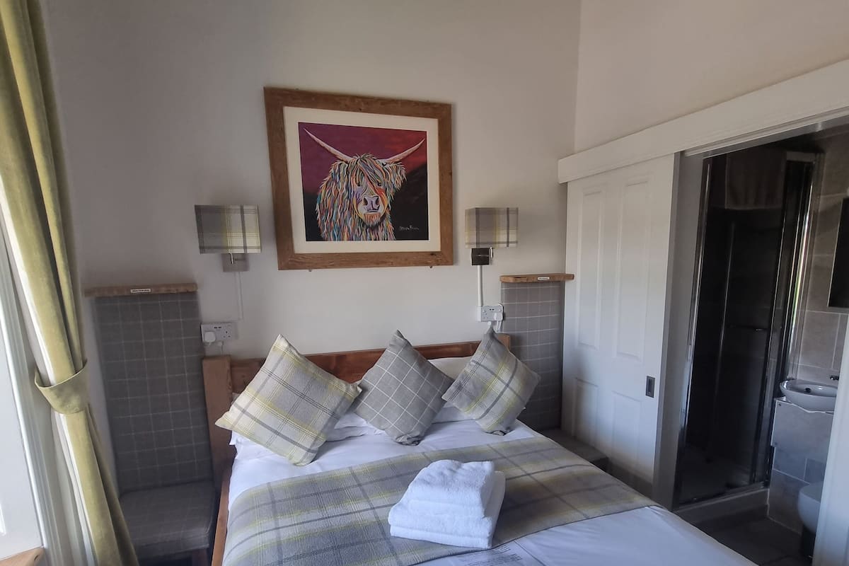 A neatly made bed with a plaid bedspread and pillows, a framed painting above it, and a bathroom visible through an open door.