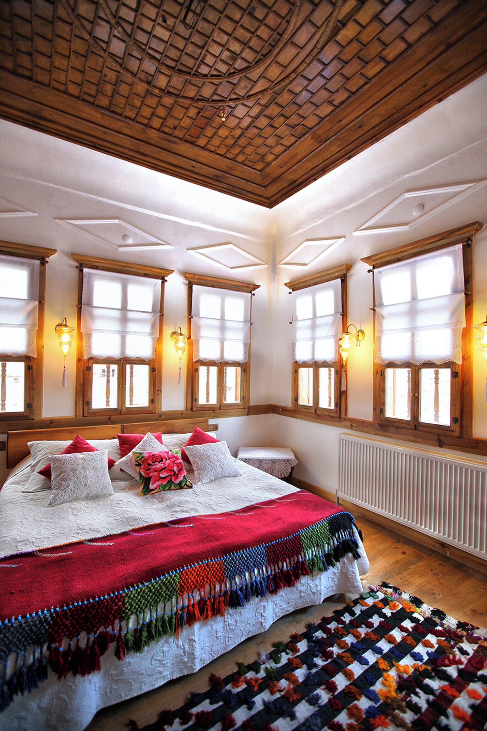 A bedroom with a wooden ceiling, large windows with curtains, a bed with pillows, and a radiator.