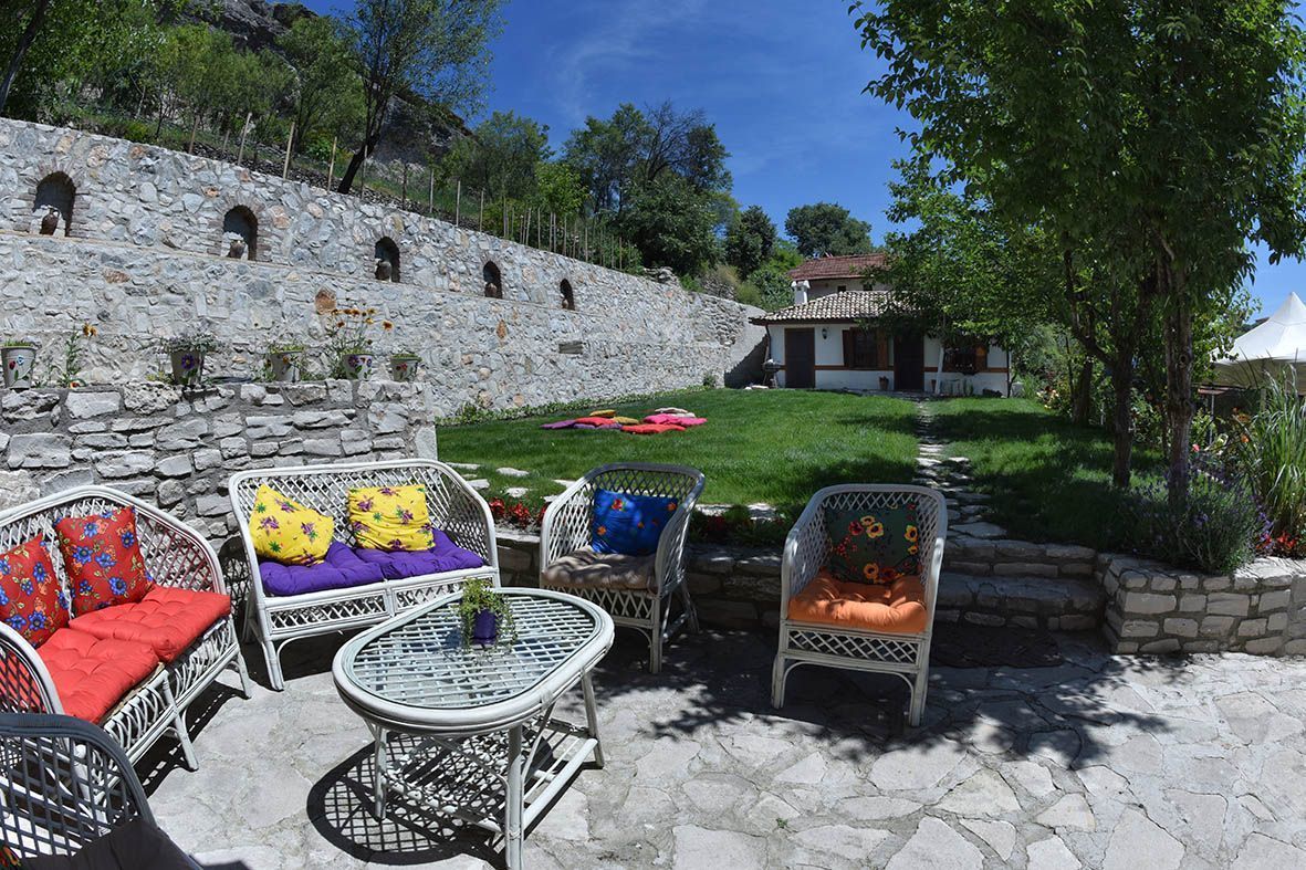 A stone wall with arched openings, a grassy area with a white building, and wicker furniture with colorful cushions.