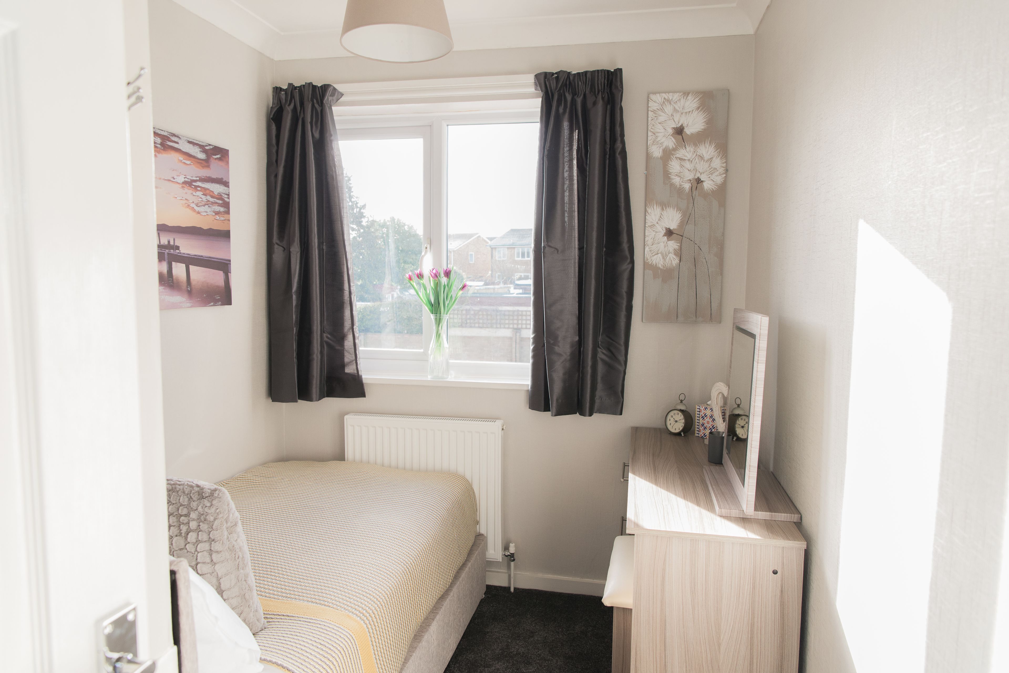 A bedroom with a bed, a dresser, a window with curtains, a radiator, a mirror, and a wall with a picture.