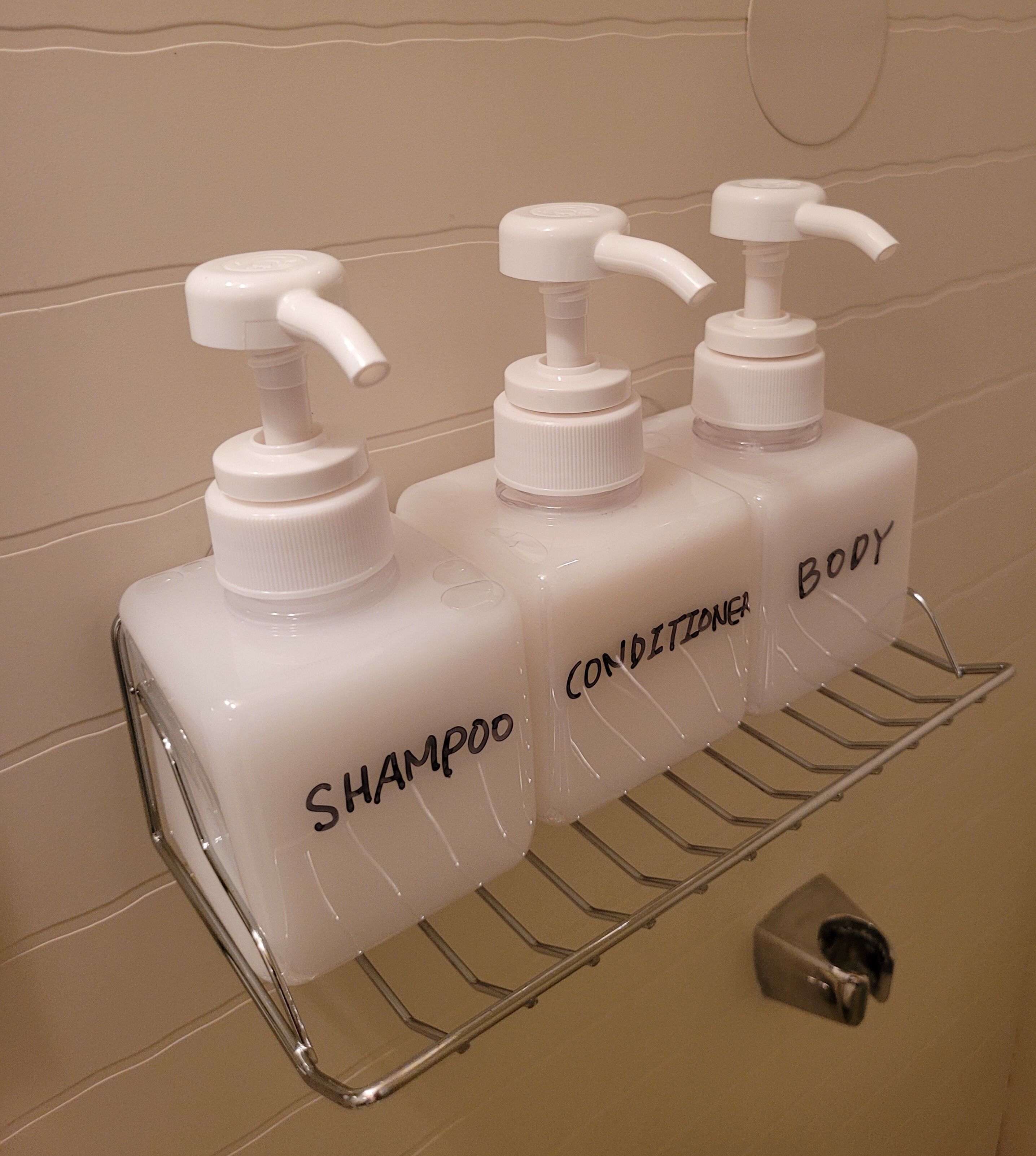 Three white bottles with pumps labeled 'SHAMPOO', 'CONDITIONER', and 'BODY' on a metal rack in a bathroom.