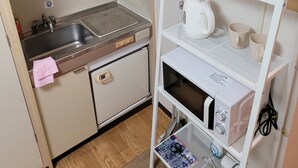 Uma cozinha compacta equipada com pia, um pequeno frigorífico, micro-ondas e uma prateleira com chávenas e chaleira.