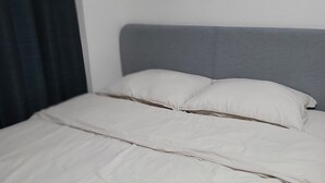 Cama bem arrumada com cabeceira cinzenta, roupa de cama branca e cortina escura parcialmente visível.