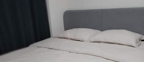 A bed with a grey headboard, white bedding, and a dark curtain to the left.
