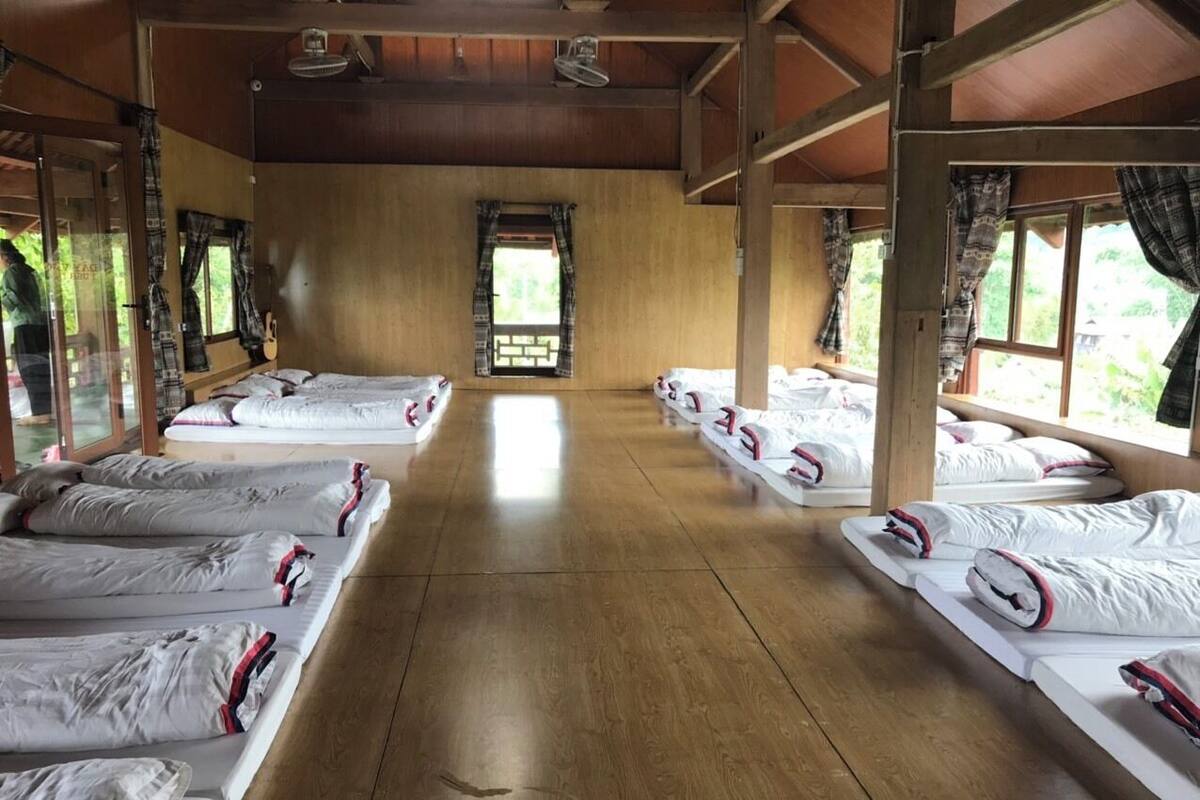 Ein Zimmer mit mehreren reihenweise angeordneten Betten, Holzböden und großen Fenstern mit Vorhängen.