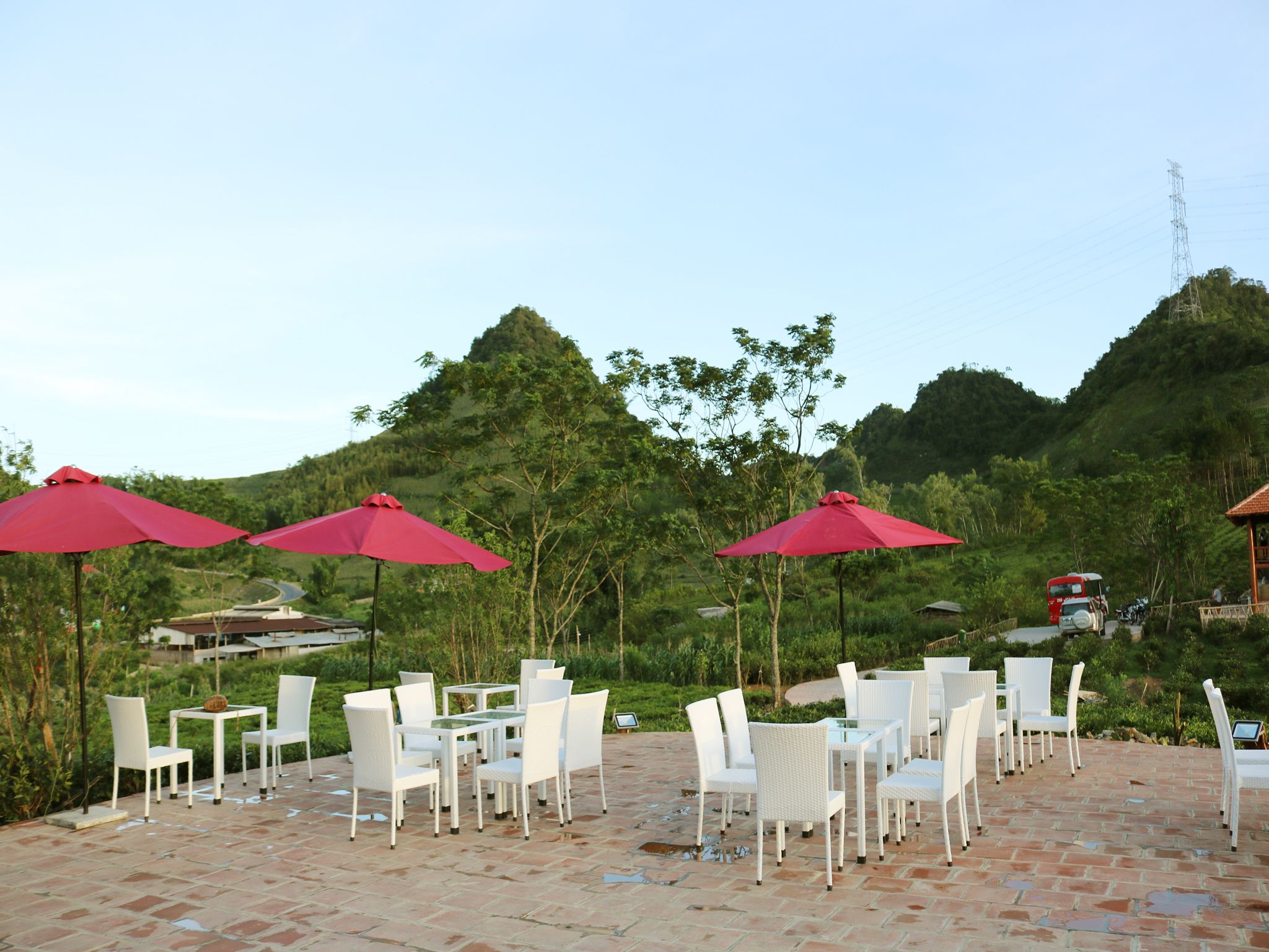 An outdoor seating area with white chairs and red umbrellas, set against a backdrop of lush greenery and hills.