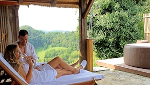 A couple relaxing on a wooden lounge chair with a bottle of champagne and a glass of wine on the table, overlooking a lush green landscape. - Vuglec Breg (Krapina)