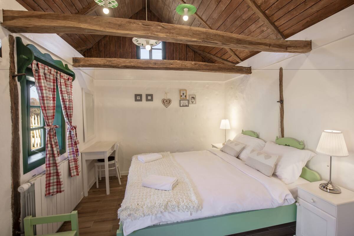A bedroom with a wooden ceiling, a bed with white bedding, a bedside table with a lamp, and a window with curtains.