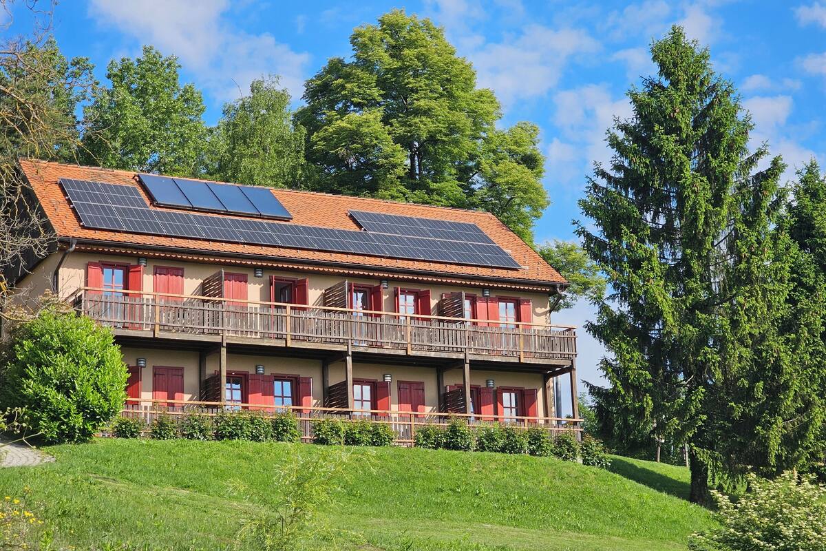A two-story building with a red-tiled roof, solar panels, and a balcony with red shutters.