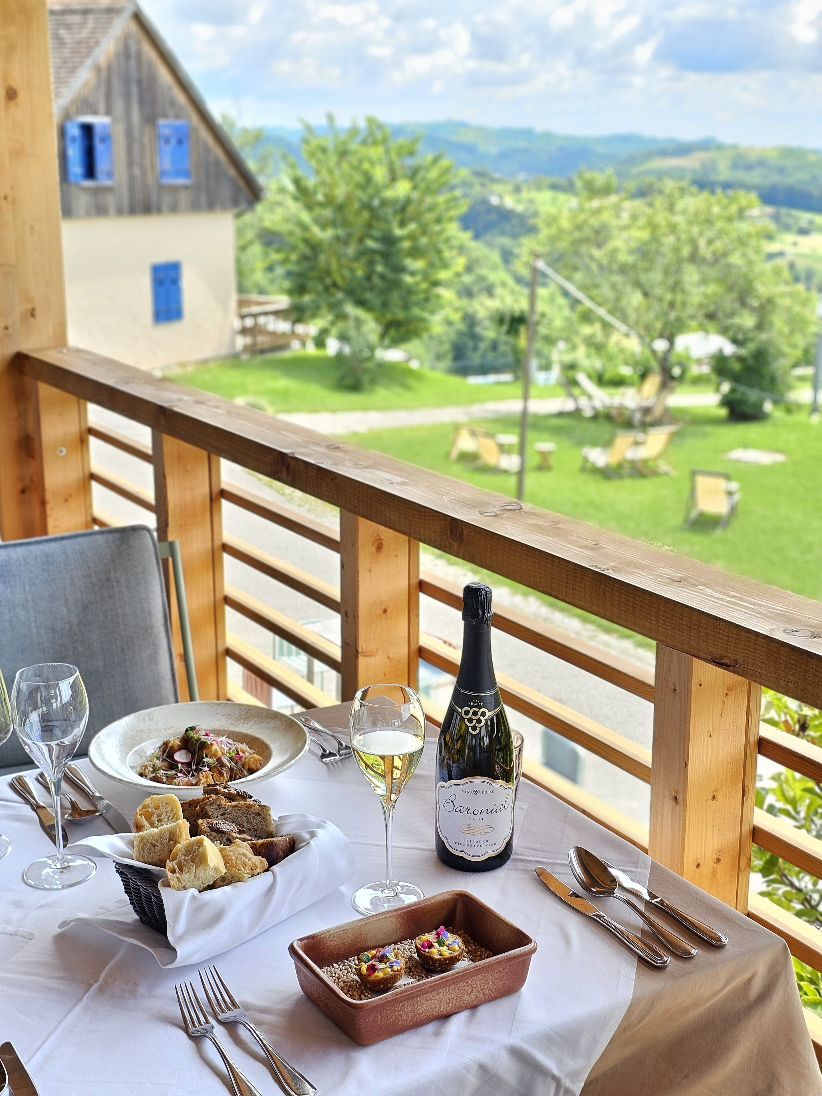 A table set for a meal with a bottle of wine, a bowl of food, and cutlery on a balcony overlooking a scenic landscape.
