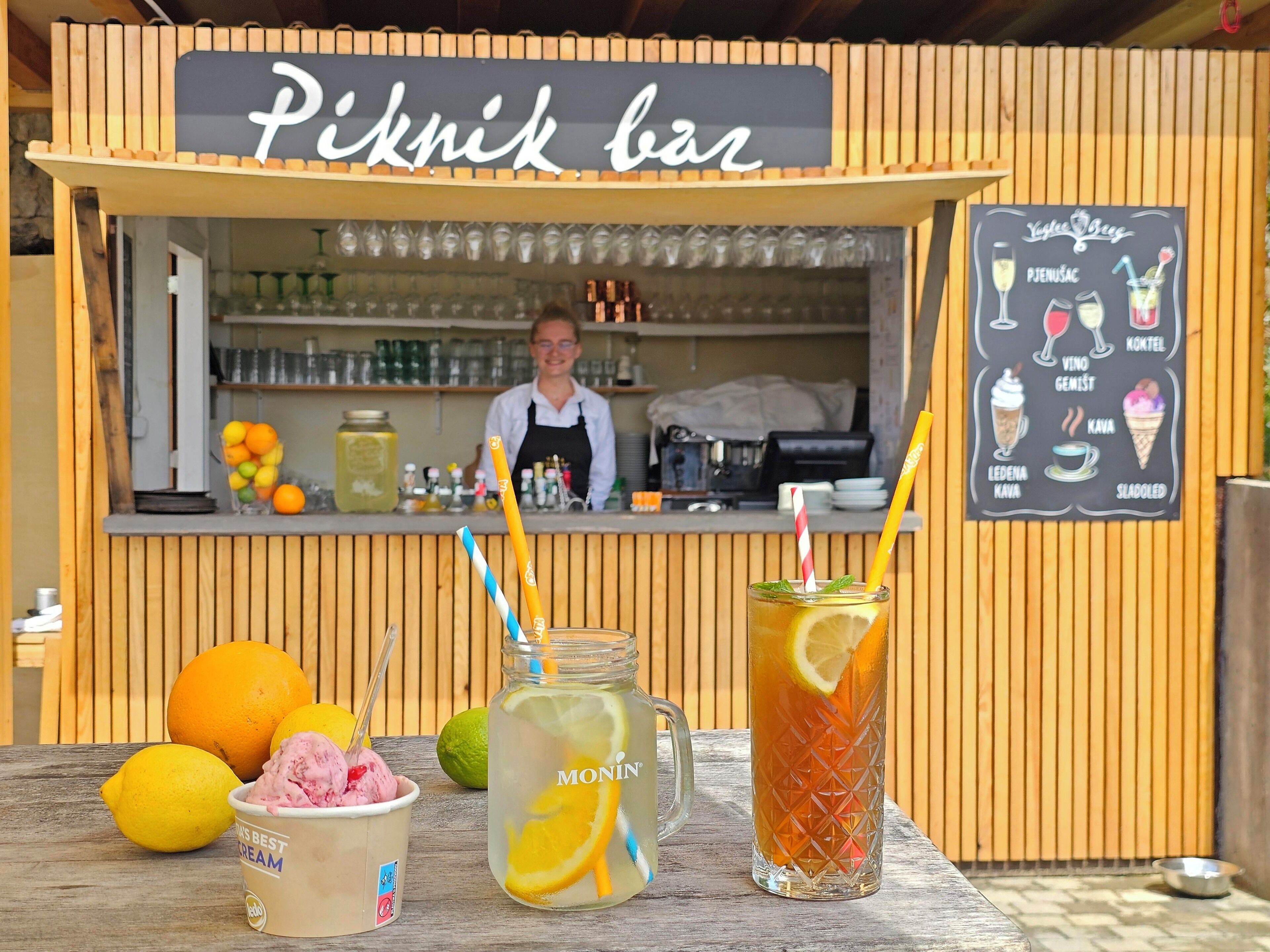 A picknick bar with a menu board, a beverage stand with drinks, and a person behind the counter.