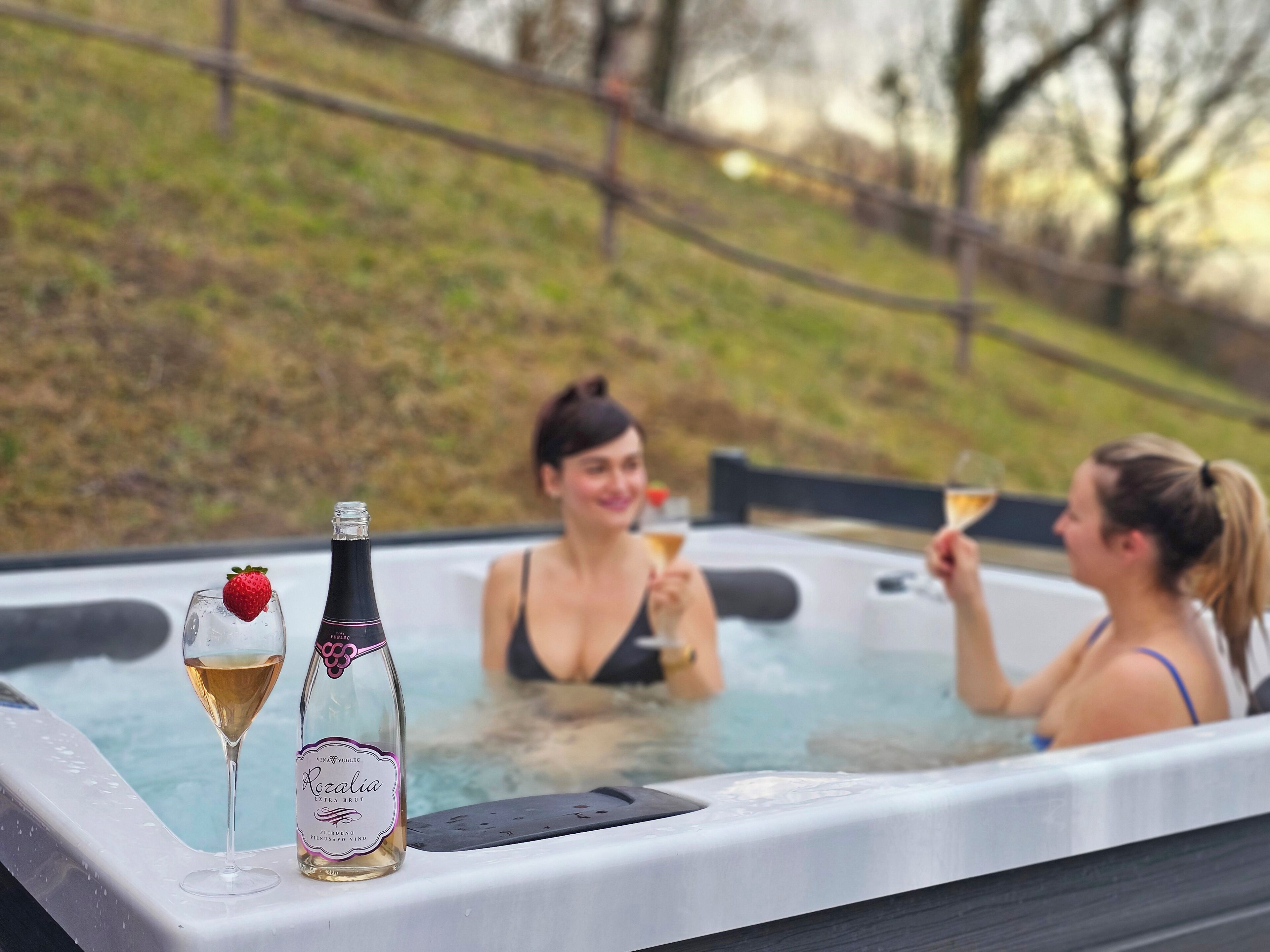 A hot tub with two people enjoying drinks, surrounded by a grassy area and trees.