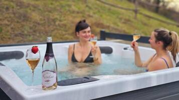 A hot tub with two people enjoying drinks, surrounded by a grassy area and trees.