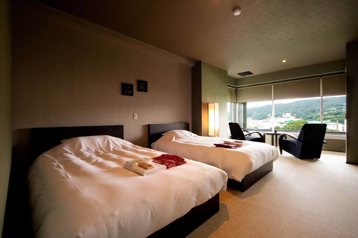 A hotel room with two beds, a large window with a view of greenery and buildings, and a chair near the window.