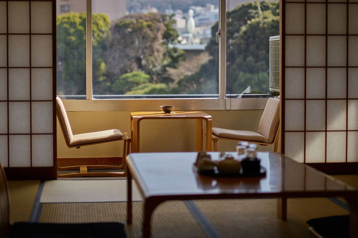 A traditional Japanese room with tatami flooring, a low table, two chairs, and a view through a large window.