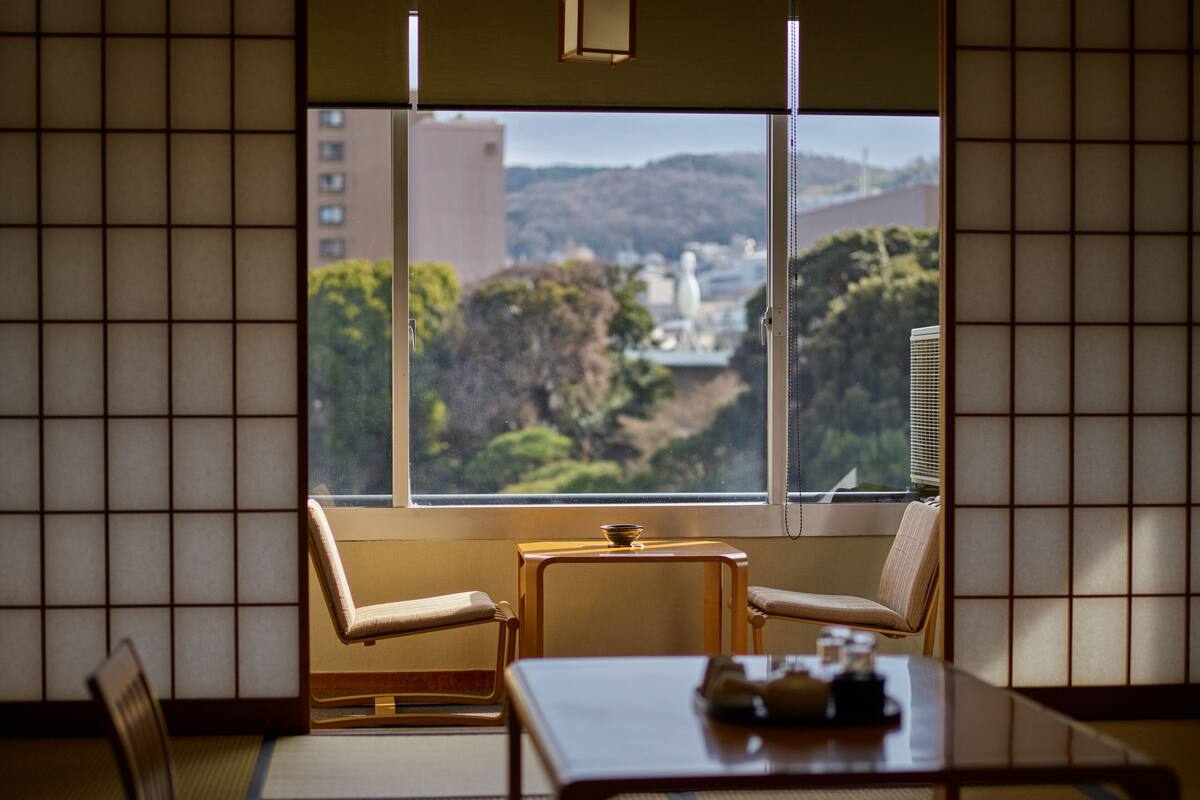 A traditional Japanese room with tatami flooring, a low table, and two chairs. A large window offers a view of a cityscape and hills.