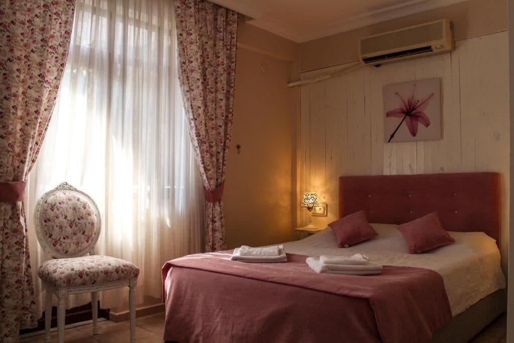 A hotel room with a bed, a chair, a wall-mounted air conditioner, and a window with curtains.