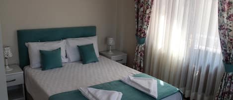 Una cama bien hecha con un cabecero color turquesa y almohadas, una mesita de noche blanca con una lámpara y cortinas florales en una ventana.