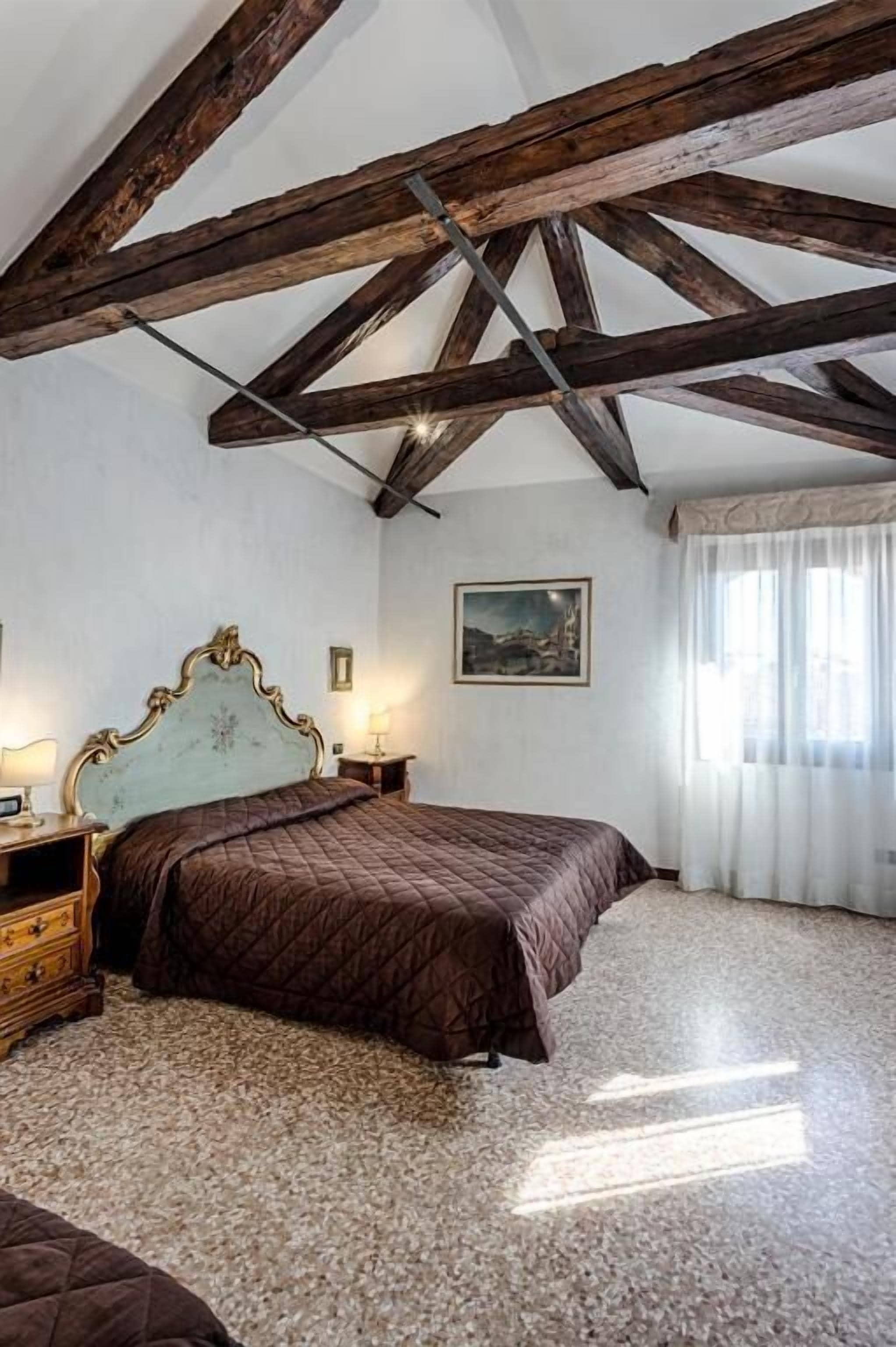 Un dormitorio con techo de madera, cama con cubrecama acolchado, mesita de noche, cómoda y un cuadro en la pared.