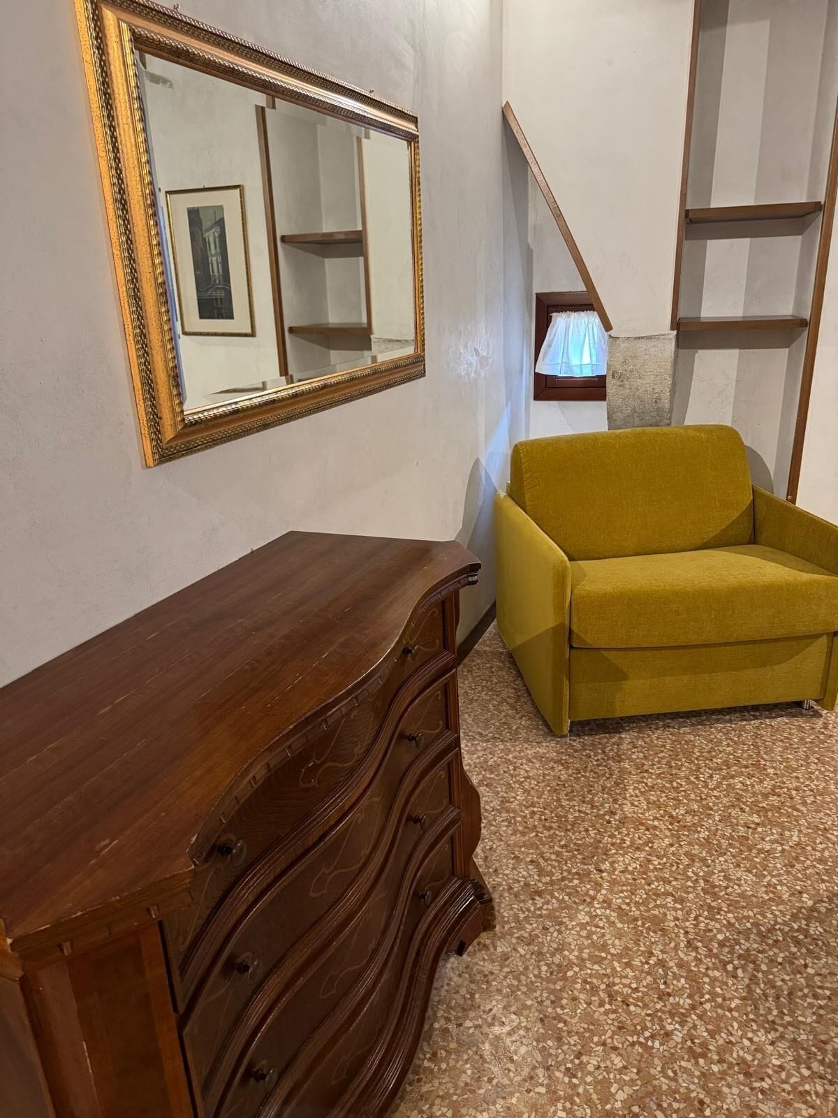 Um quarto com cômoda de madeira, sofá amarelo mostarda, um espelho grande e uma escada.