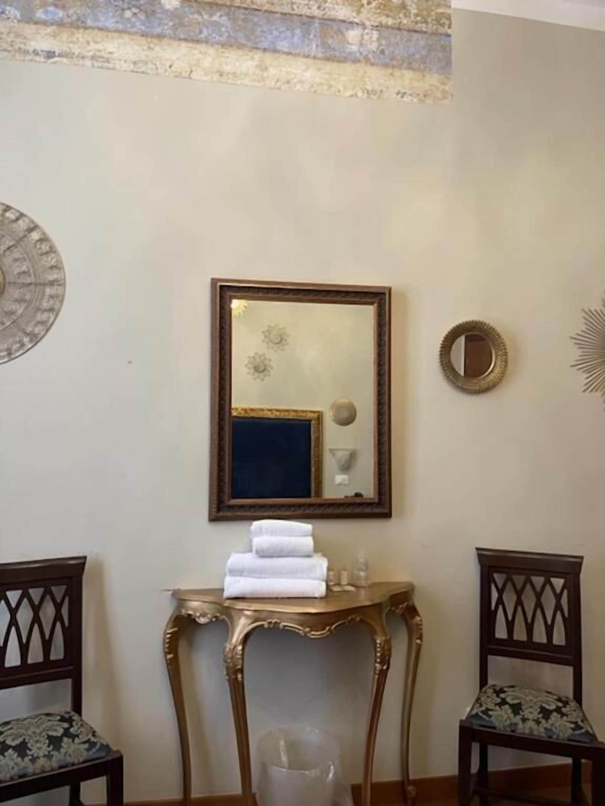 A room with a large mirror, a small table with towels, two chairs, and decorative wall art.