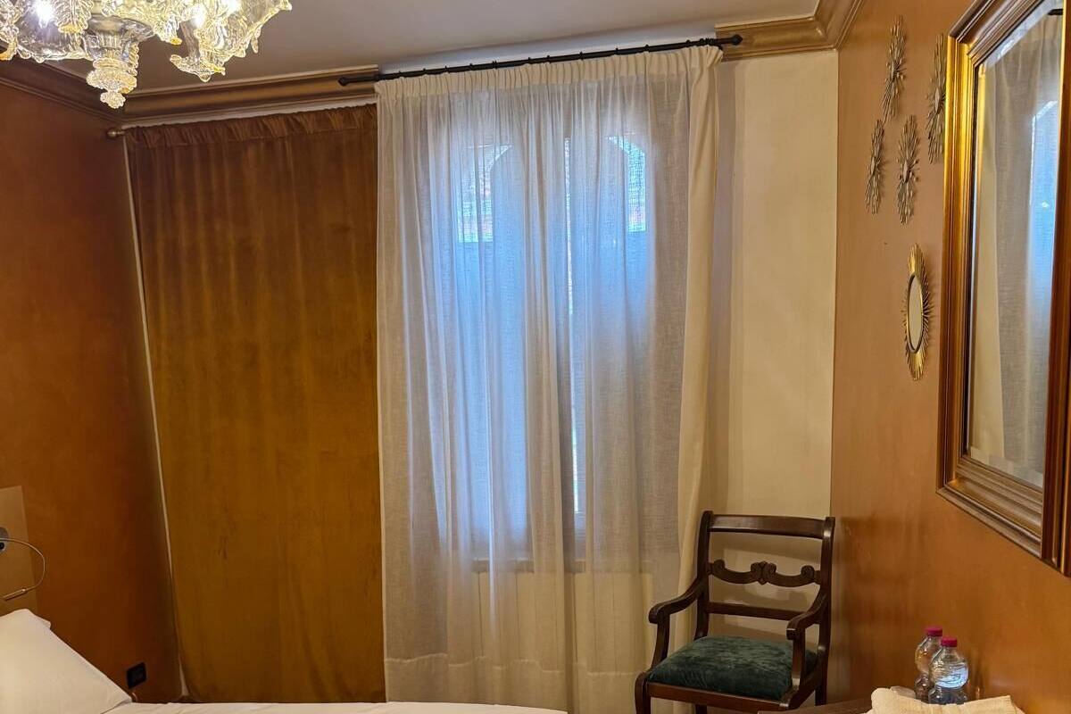 A hotel room with a large bed, a wooden chair, a small table with towels, a trash bin, and a chandelier.