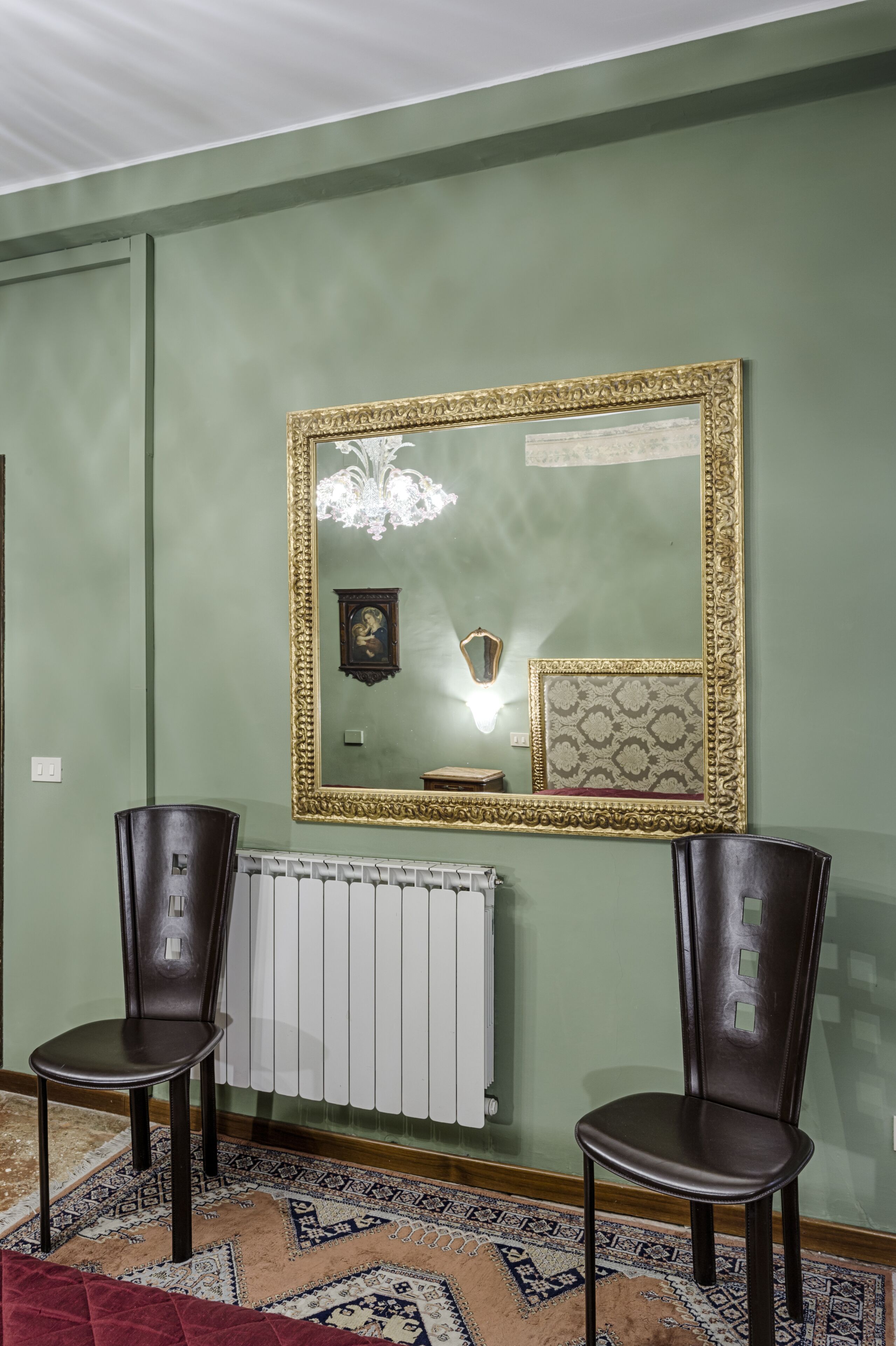 A room with a large ornate mirror, two chairs, a radiator, and a patterned rug.
