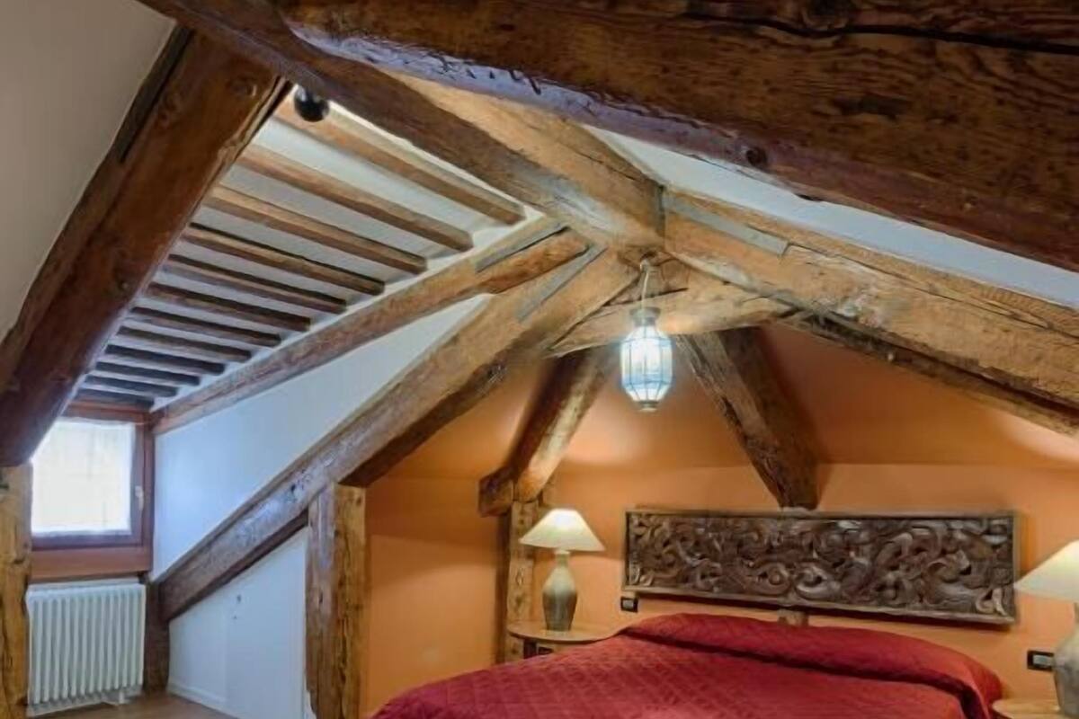 A bedroom with a sloped ceiling, wooden beams, a red bedspread, and a nightstand with a lamp.