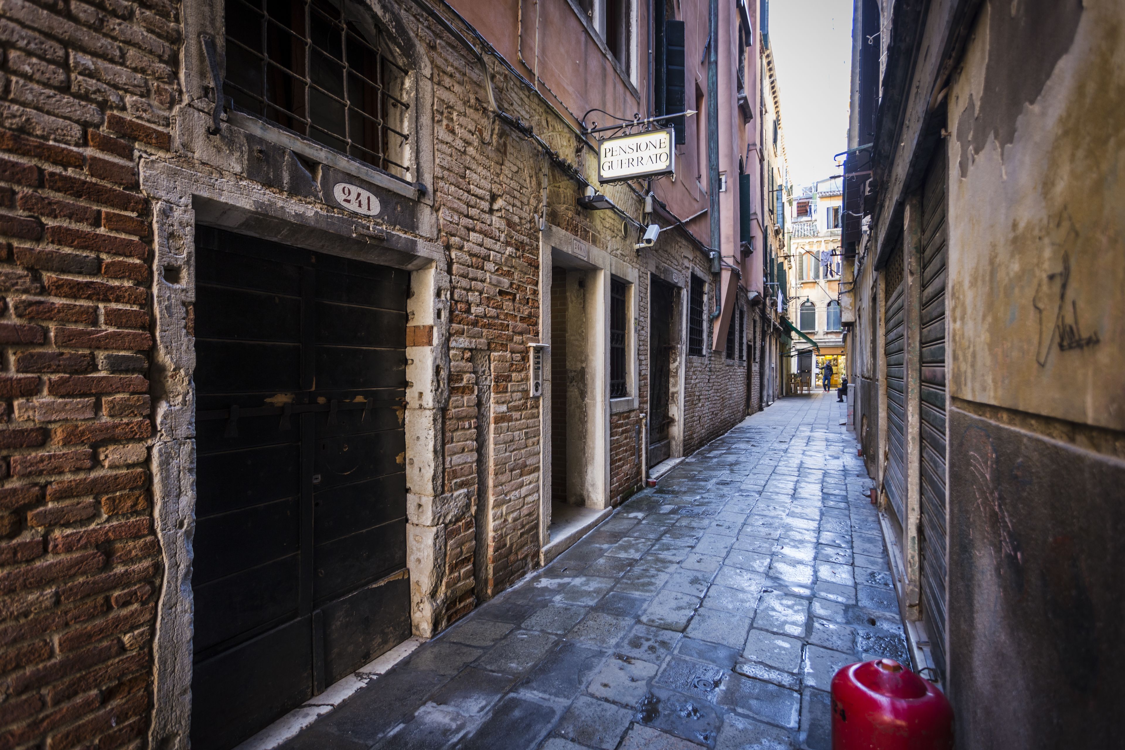 A narrow alleyway with brick buildings, a closed metal gate, and a red object on the ground.