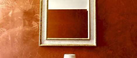 Un miroir encadré est accroché à un mur à texture, et une lampe est posée sur une petite table en contrebas.