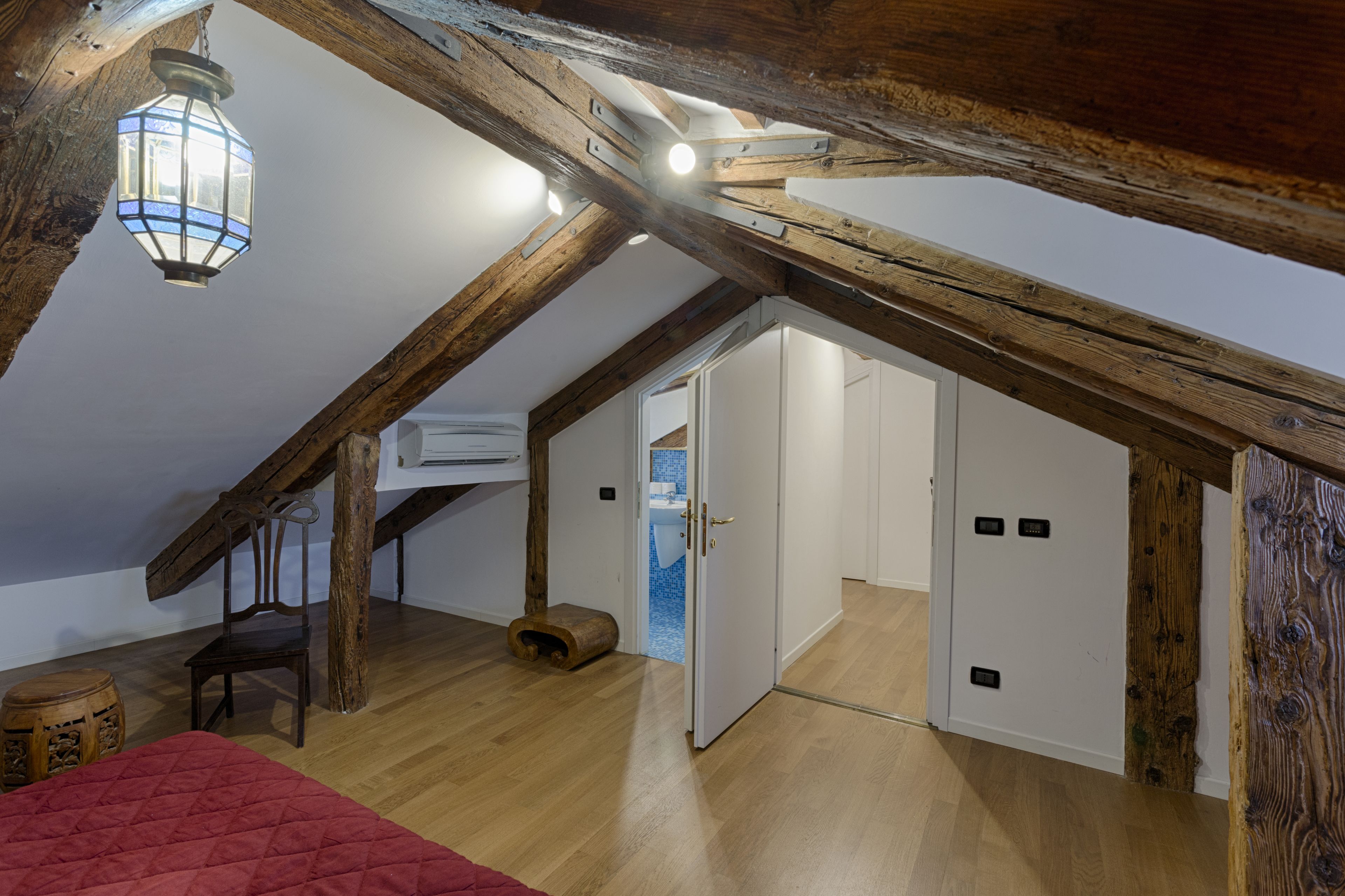 A room with exposed wooden beams, a hanging lantern, a red bed, and a wooden chair.