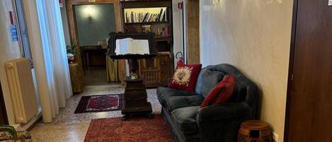 A living room with a dark sofa, red cushions, a wooden side table, a floor lamp, a chandelier, a bookshelf, and a small round table.