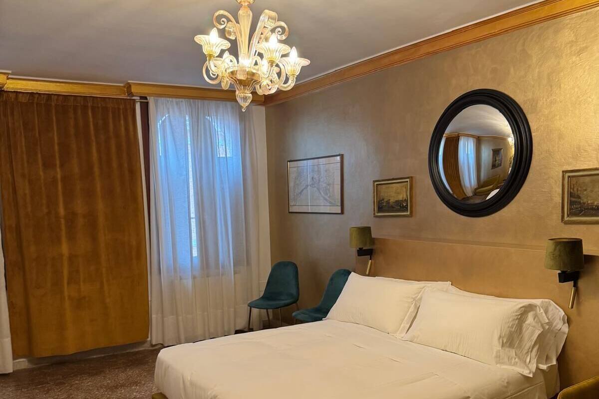A hotel room with a large bed, a chandelier, a round mirror, and a window with curtains.