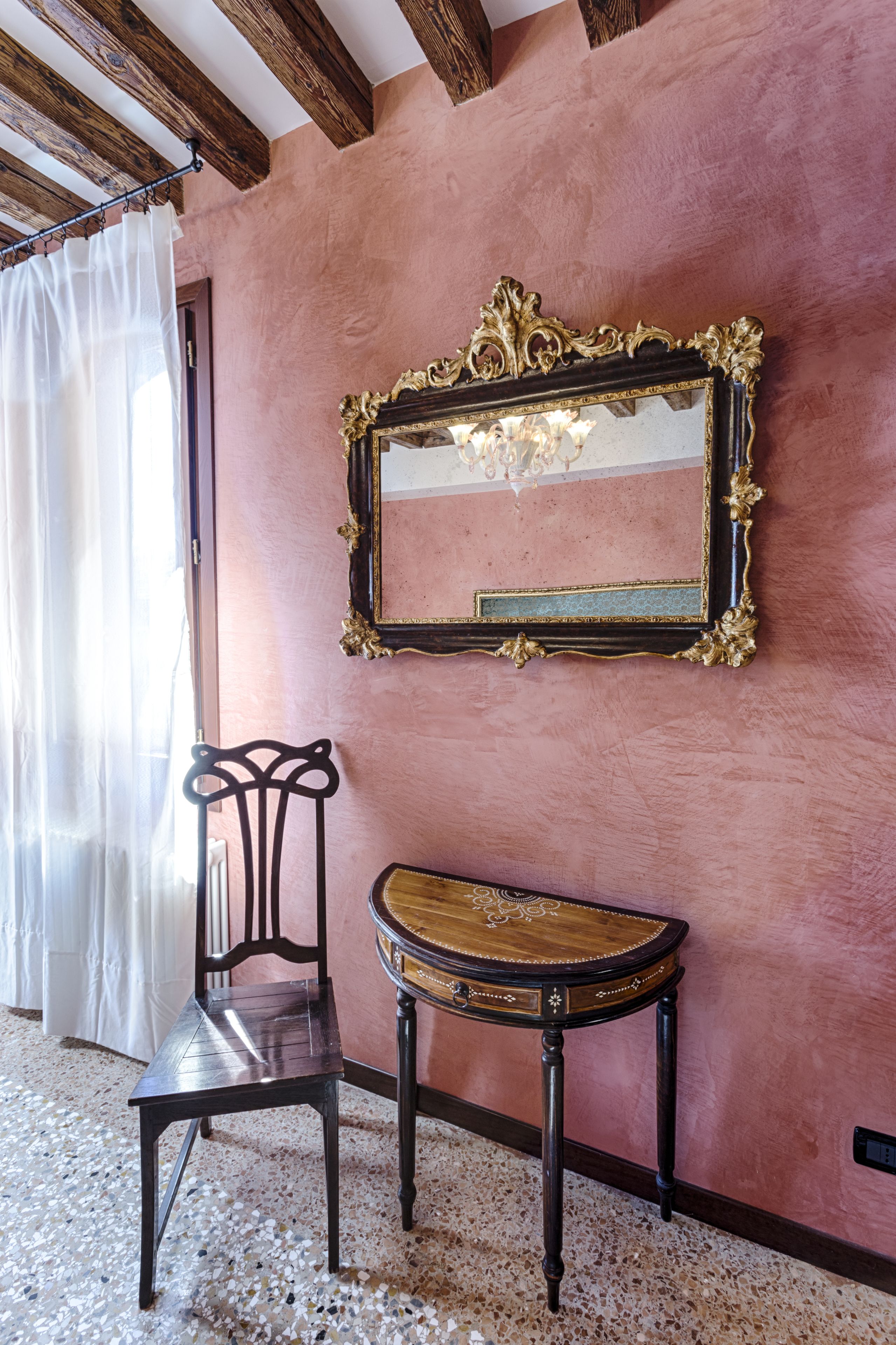Una habitación con una pared rosa, una silla de madera, una mesita pequeña y un espejo con un marco decorado.