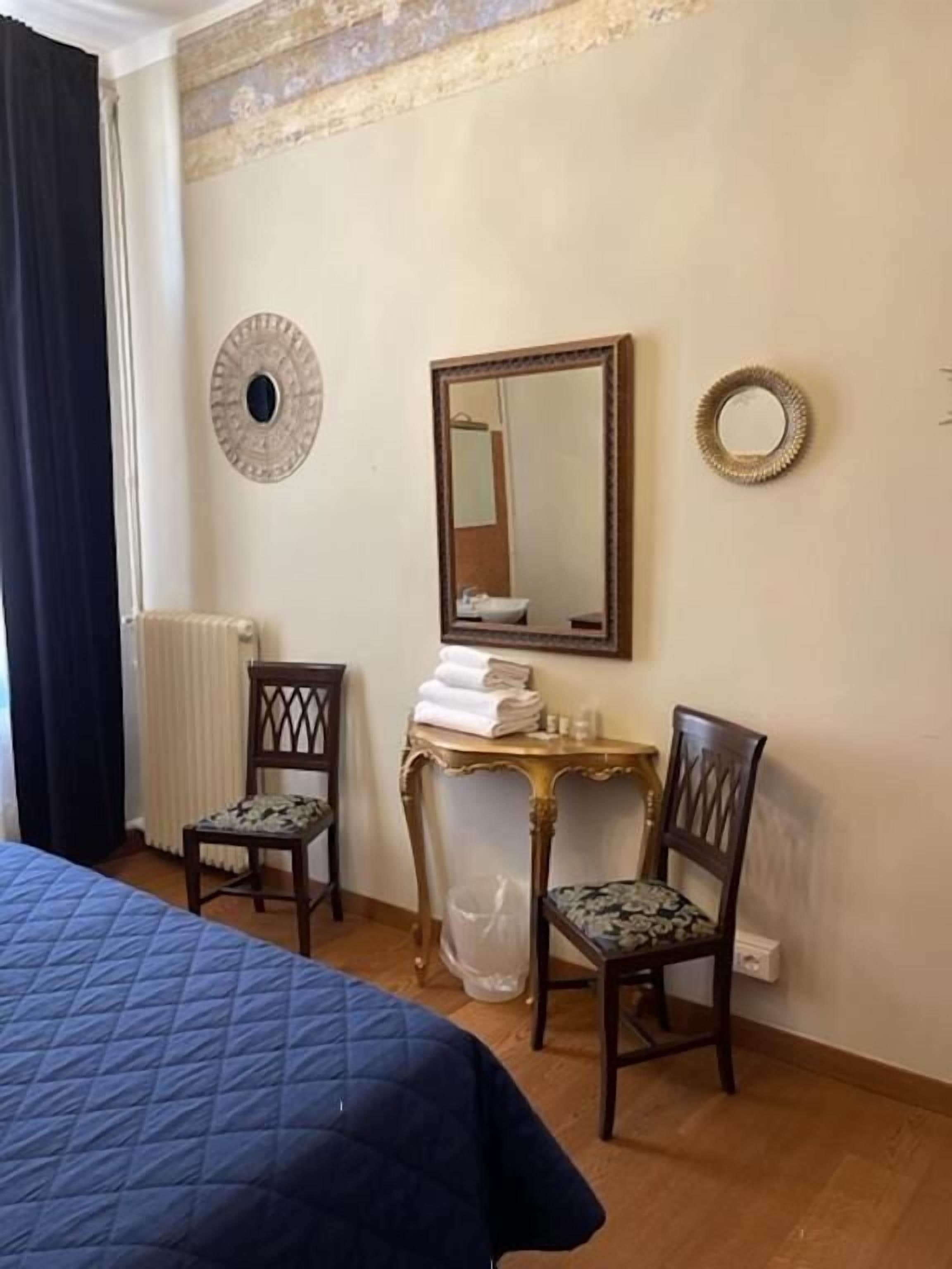 A hotel room with a bed, two chairs, a small table with towels, a mirror, and a radiator.