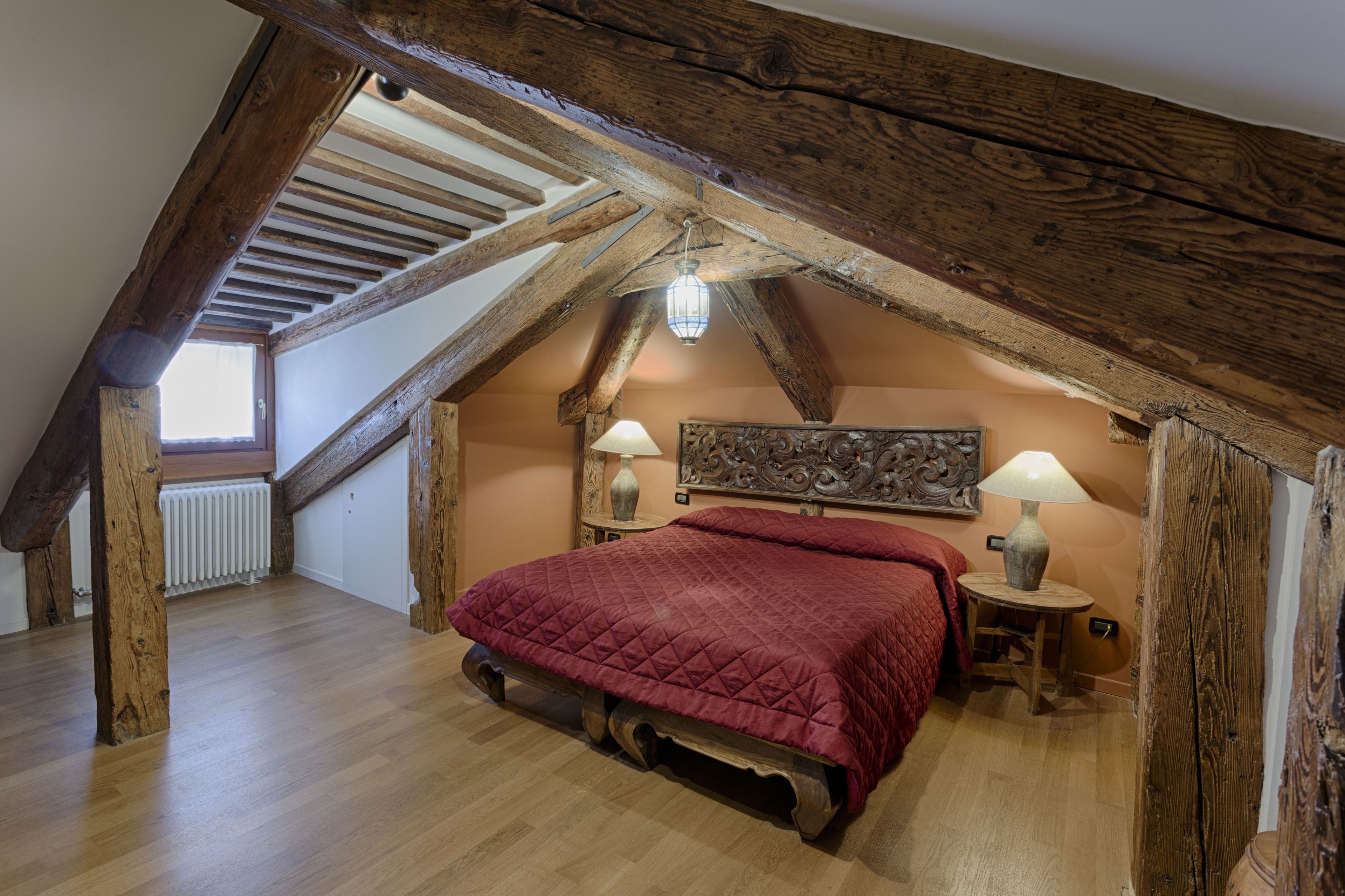 Un dormitorio con techo inclinado, vigas de madera, una cama con colcha roja, dos lámparas de noche y un radiador.