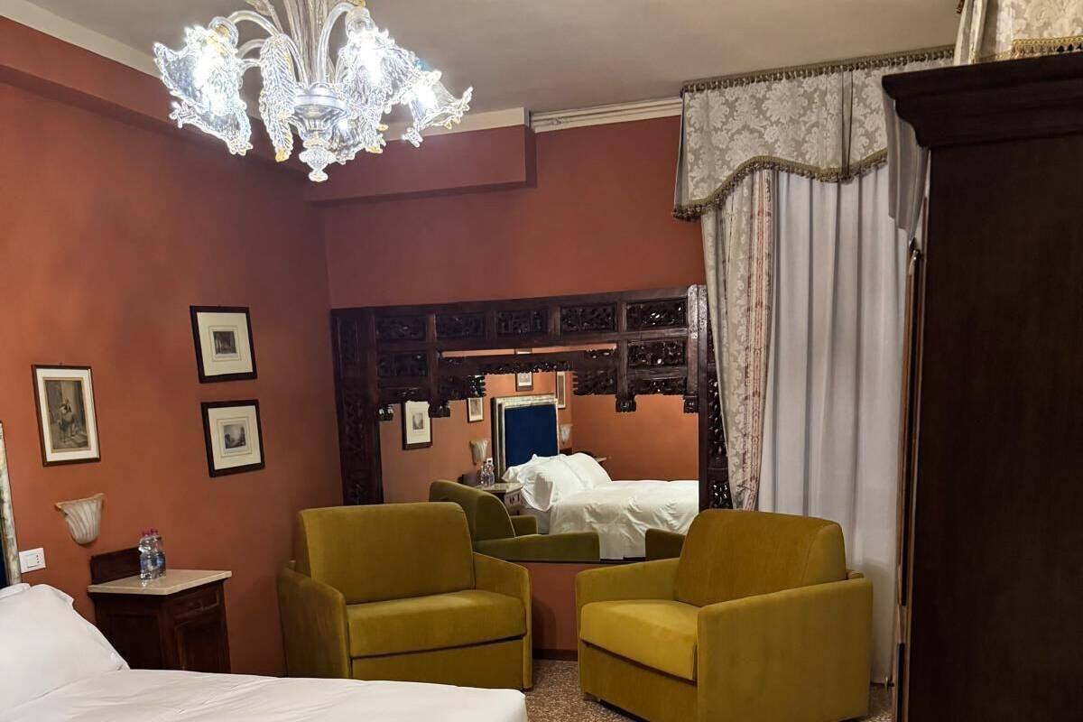 A hotel room with a bed, two yellow armchairs, a wooden bench, a chandelier, and a mirror with a blue frame.