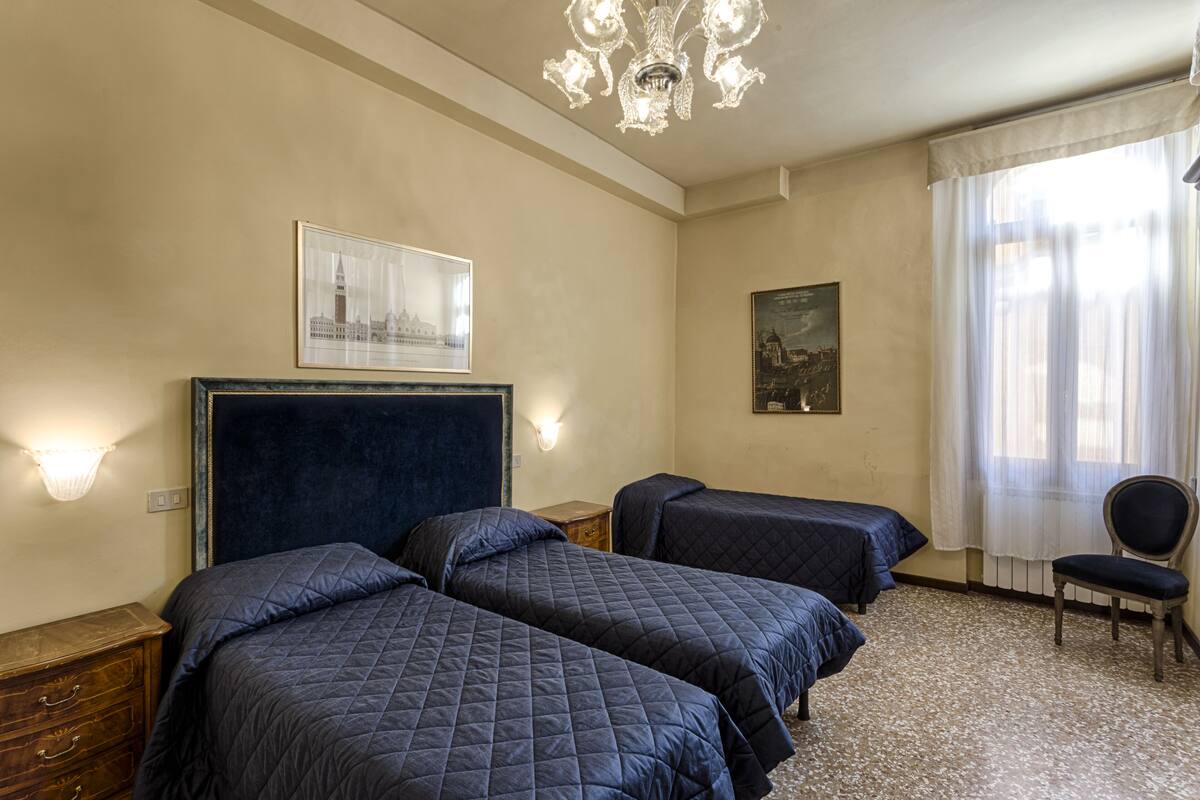 A hotel room with two beds, a chair, a chandelier, and two wall-mounted lamps.