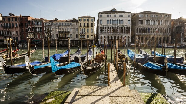 Un canal con góndolas amarradas a muelles de madera, rodeado de edificios históricos con ventanas arqueadas y balcones.