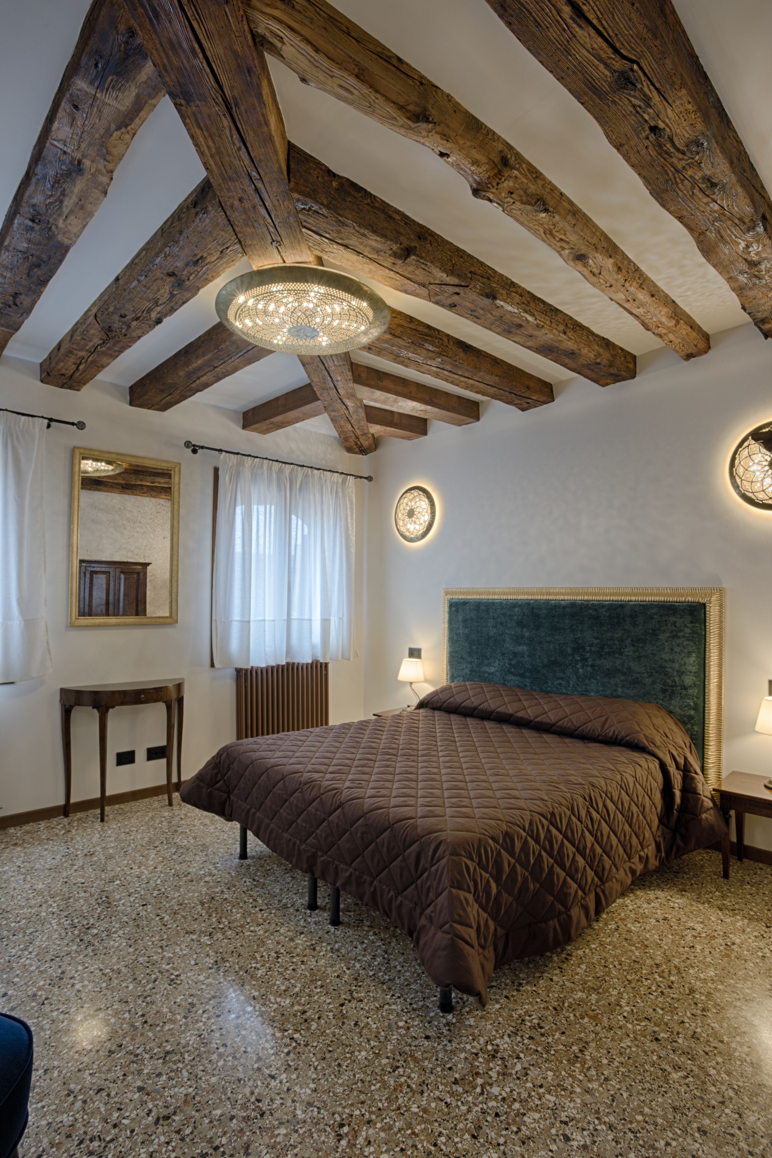 A bedroom with a large bed, wooden beams, a chandelier, a mirror, and a round wall clock.