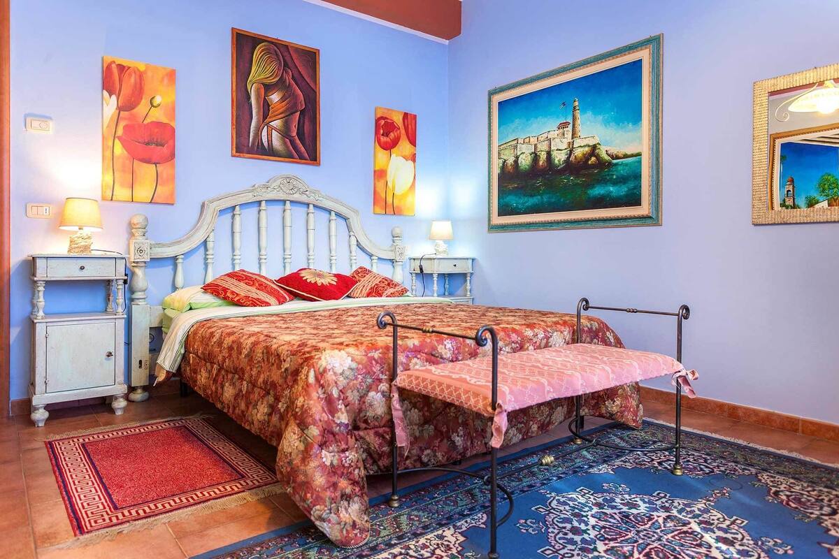 Ein Schlafzimmer mit einem Bett, zwei Nachttischen, einem Beistelltisch, einem roten Teppich und einem großen Gemälde eines Leuchtturms.