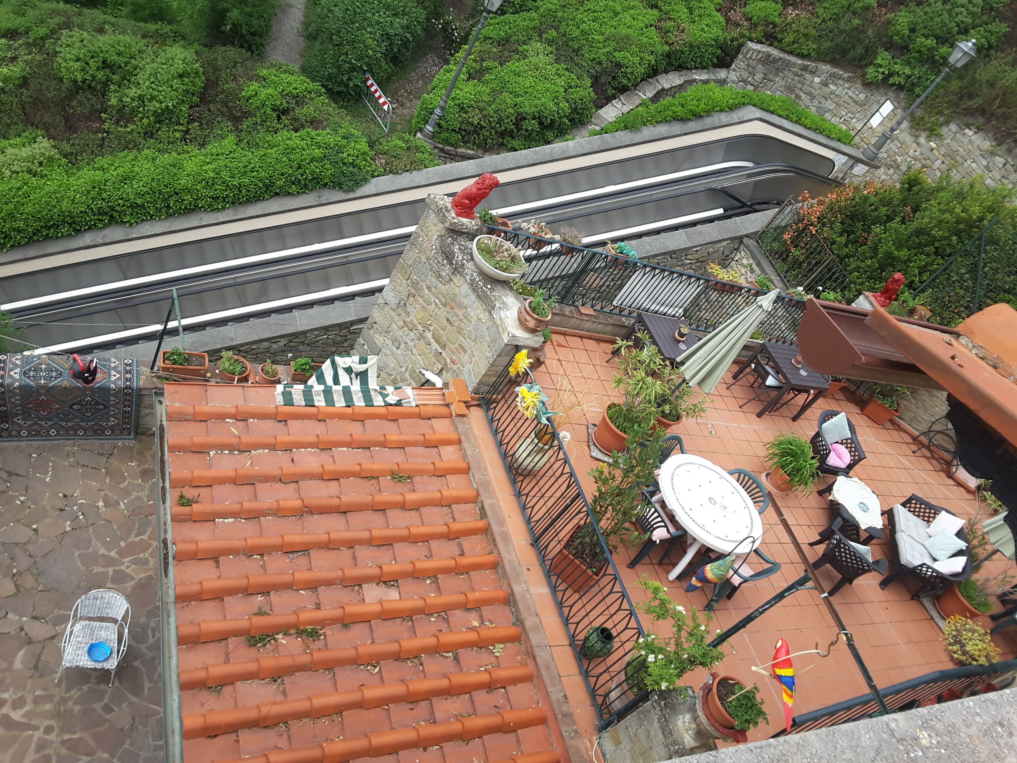 A rooftop terrace with terracotta tiles, potted plants, outdoor furniture, and a view of a road and greenery.