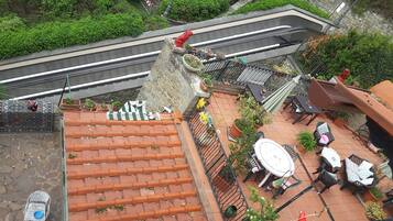 A rooftop terrace with terracotta tiles, potted plants, outdoor furniture, and a view of a road and greenery.